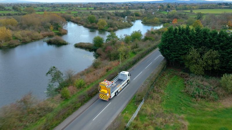 Drone view of a white impact protection vehicle travelling along a rural highway beside a lake.